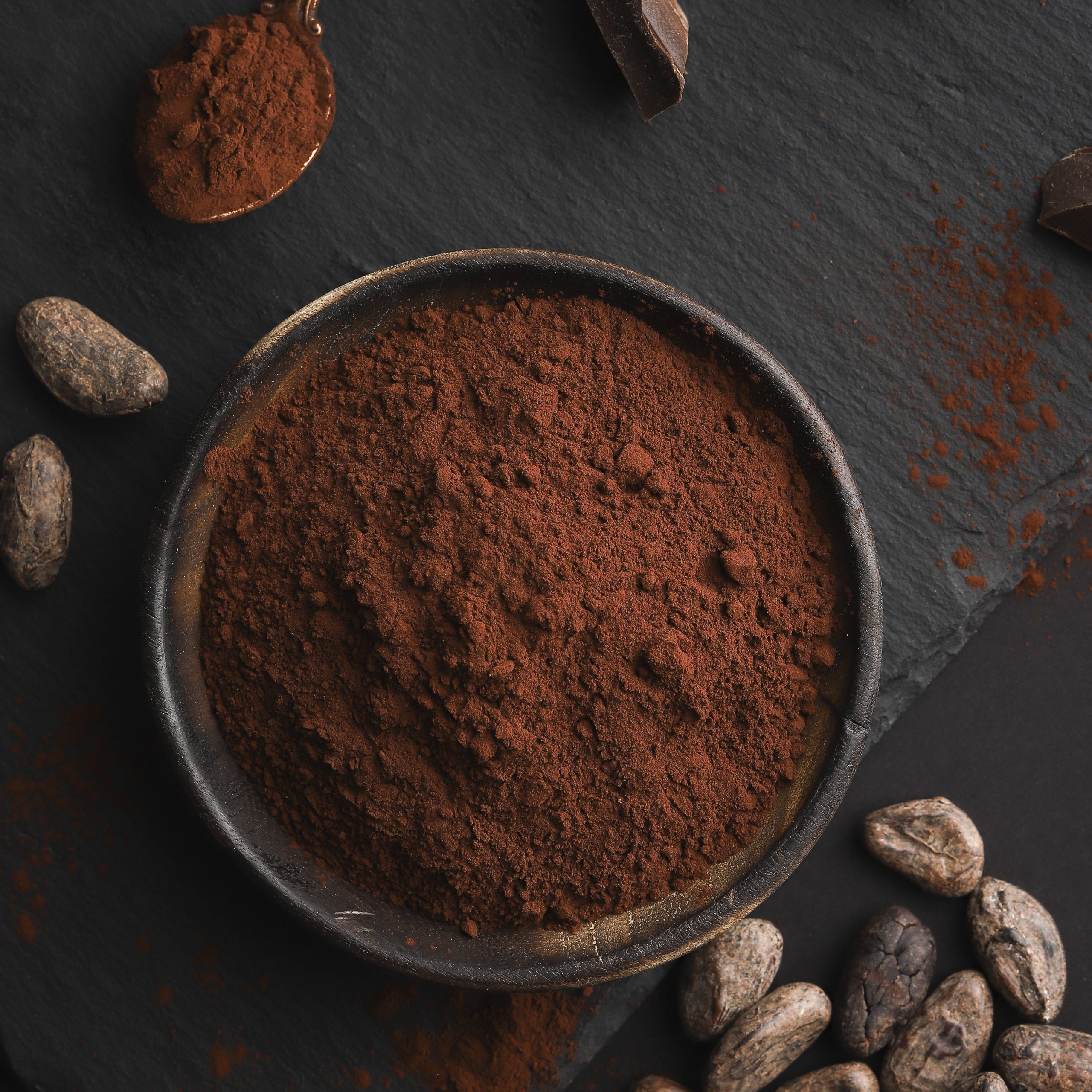 Cocoa powder in a wooden bowl. Top view. Flat lay. Broken chocolate nuts pieces, cocoa bens and cocoa powder on dark stone background.